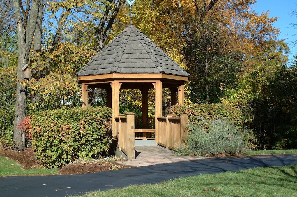 Large wooden gazebo off a walking path set within shrubs and trees.