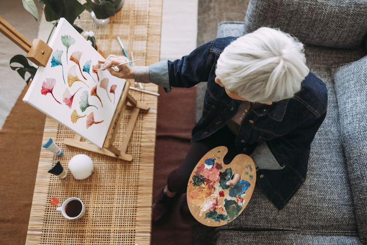 senior woman painting on a campus on her couch at home
